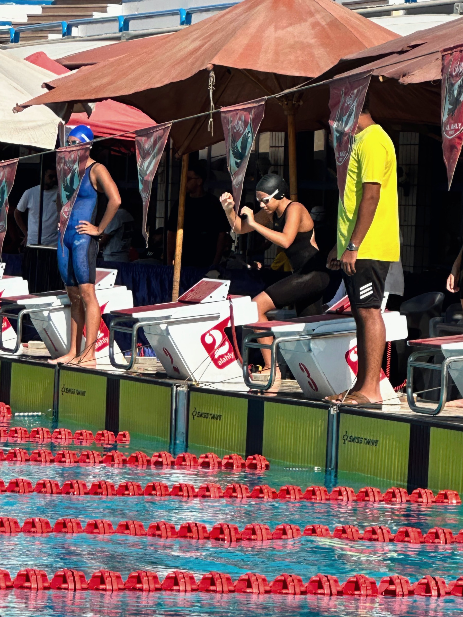 Swimmers behind the blocks before their heat, one swinging his arms, officials watching