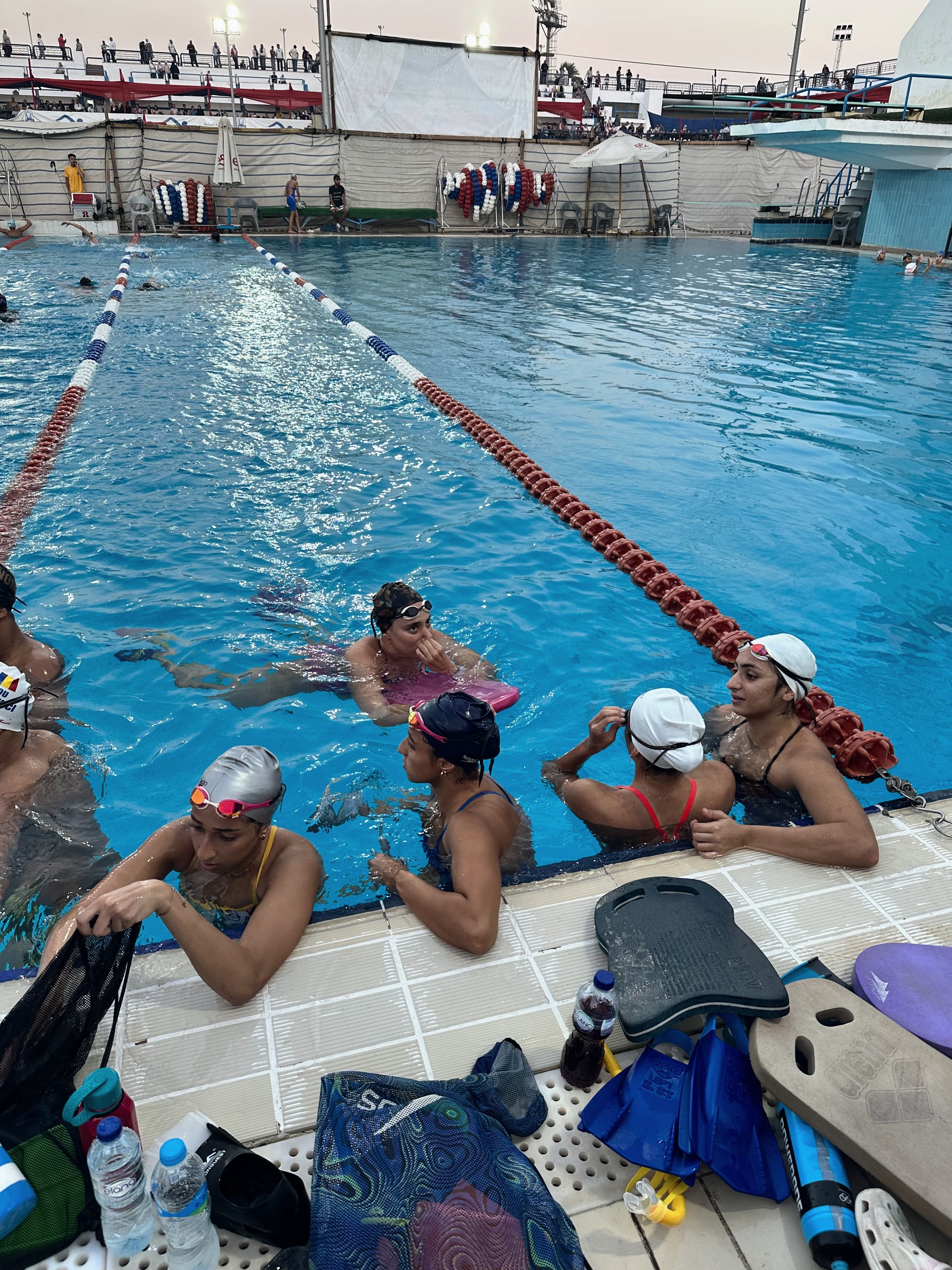 An evening session: swimmers resting at the wall, kickboards, fins and a snorkel on the deck